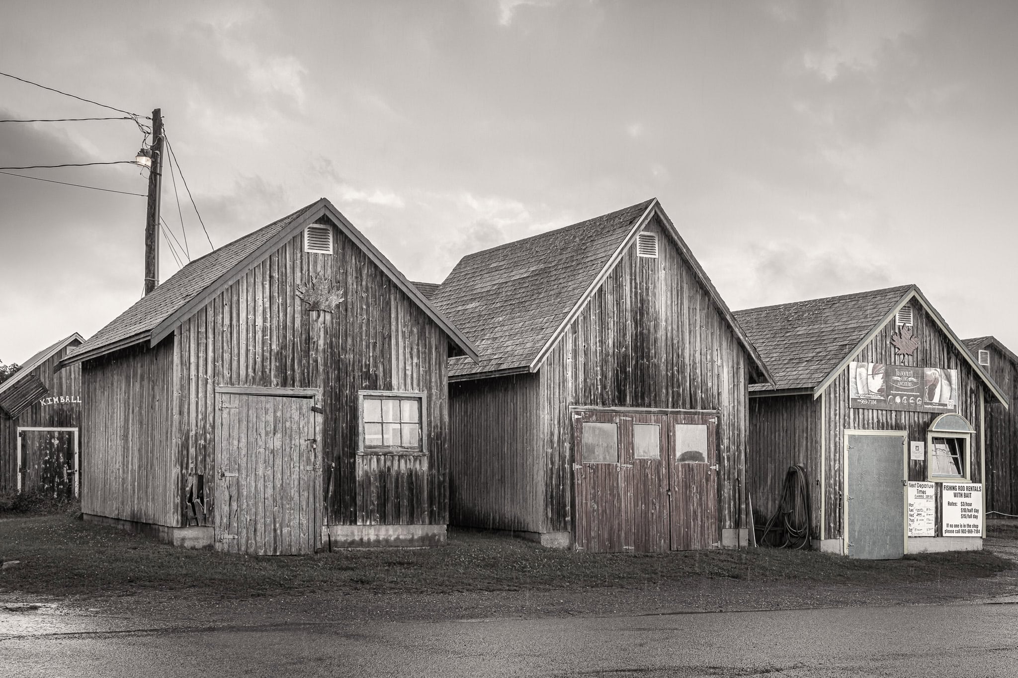Row of weathered wooden fishing huts on a rainy shoreline in Prince Edward Island, Canada, with signs, textures, and grey coastal light.